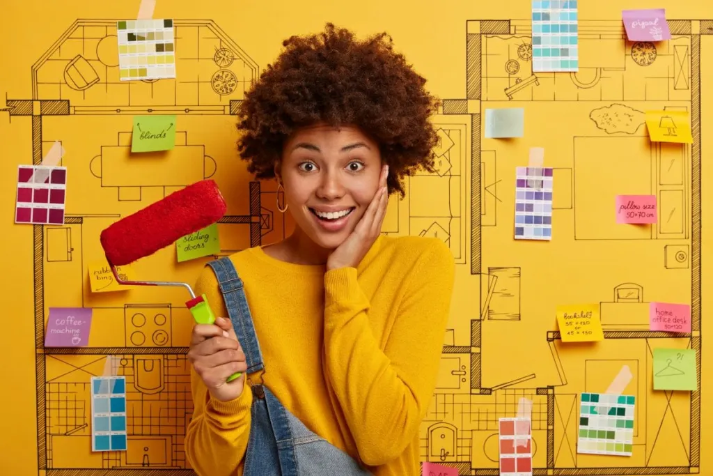 Smiling woman holding a paint roller in front of a yellow wall covered with colour swatches